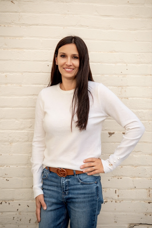 Woman wearing a white long-sleeve shirt and blue jeans against a light wooden paneled wall.