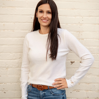 Woman wearing a white long-sleeve shirt and blue jeans against a light wooden paneled wall.
